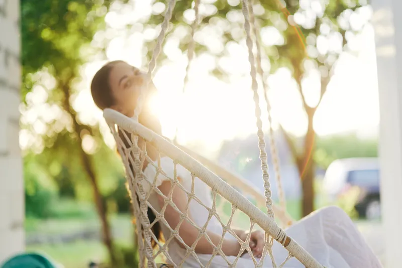 Young woman in a white dress relaxing in a swing with sunlight shining in a serene garden, symbolizing tranquility and happiness in nature