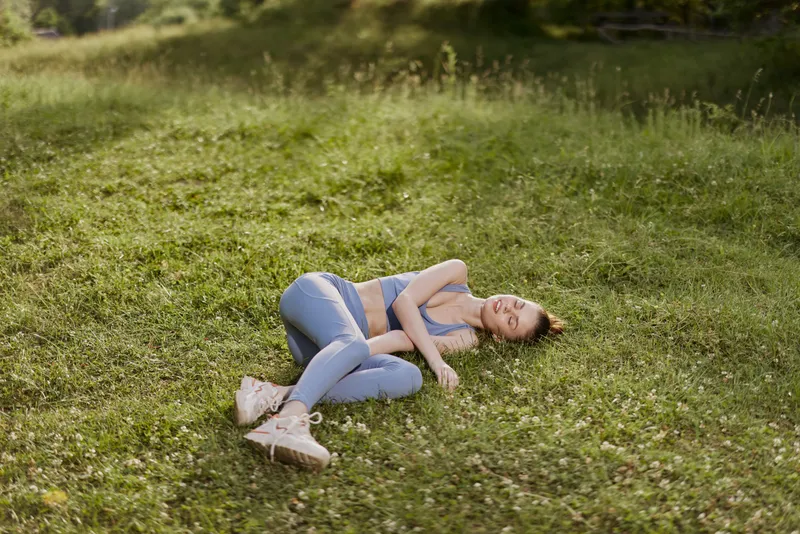 Young woman in activewear lying on green grass, embodying relaxation and fitness in a serene outdoor setting, promoting health and wellness
