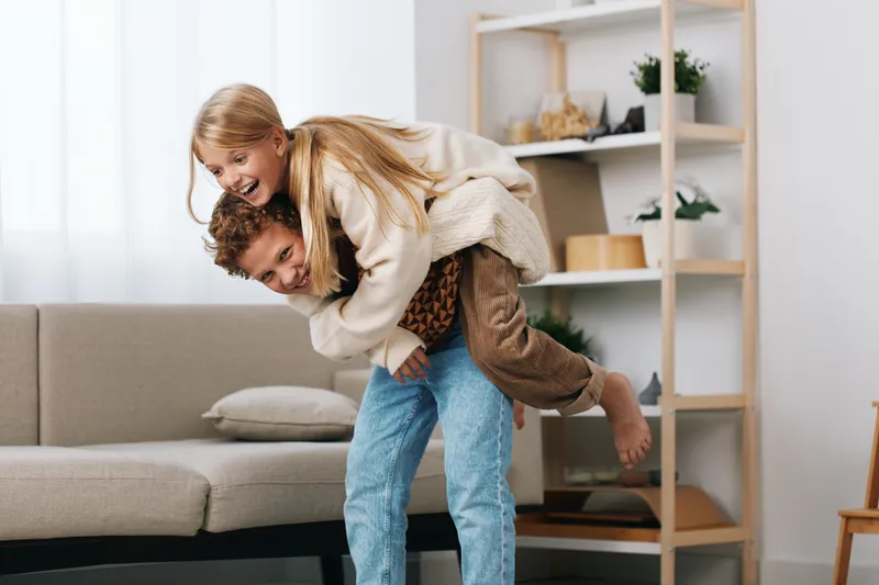Authentic playful moment of a couple sharing a piggyback indoors, capturing candid lifestyle and everyday joy in a cozy living room with soft light, warm tones, and genuine connection.