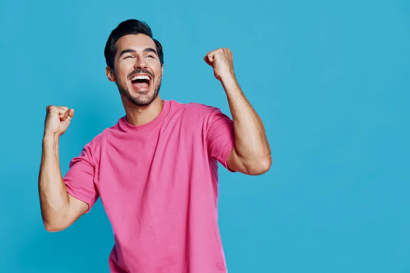 Full body joyful cheerful young man wearing pink t-shirt celebrating success with raised hands against a bright blue background. Positive emotions concept.