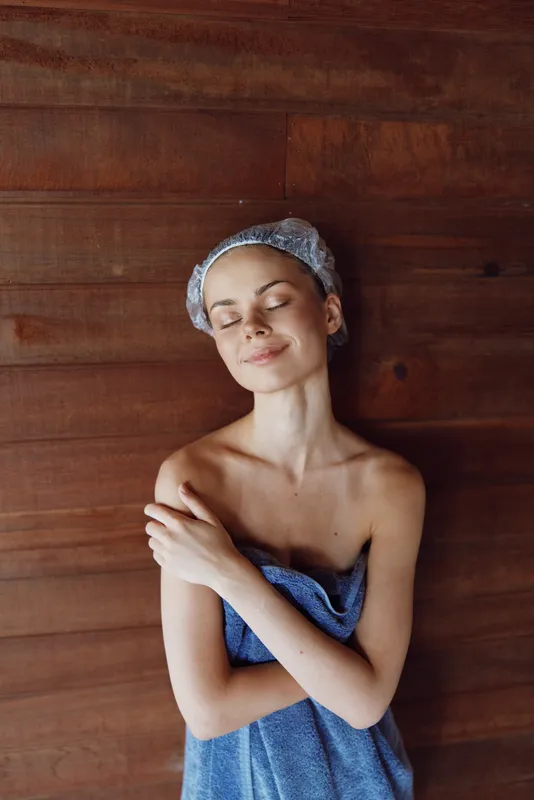 relaxed woman wrapped in blue towel with hairnet stands against wooden wall, eyes closed, enjoying calm moment of wellness and self-care in warm indoor spa environment