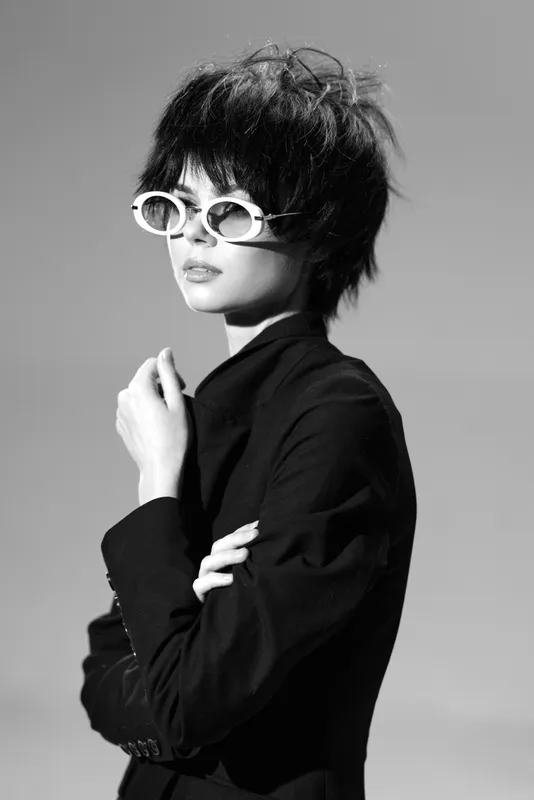 Black and white portrait of a stylish woman with short hair and white sunglasses wearing a dark jacket.