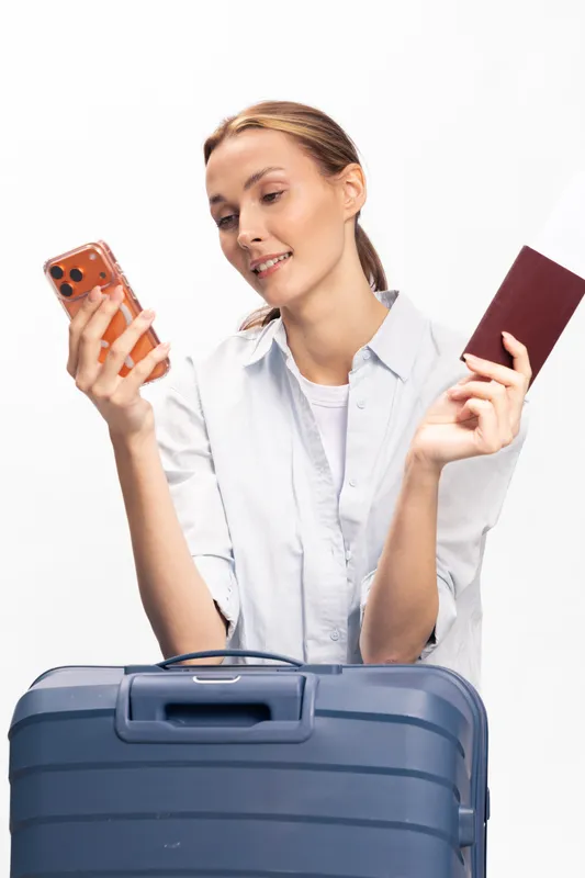 Young woman holding passport and smartphone looks at screen smiling. Blue suitcase in front. Concept of travel planning, tourism, and vacation preparation on white background.