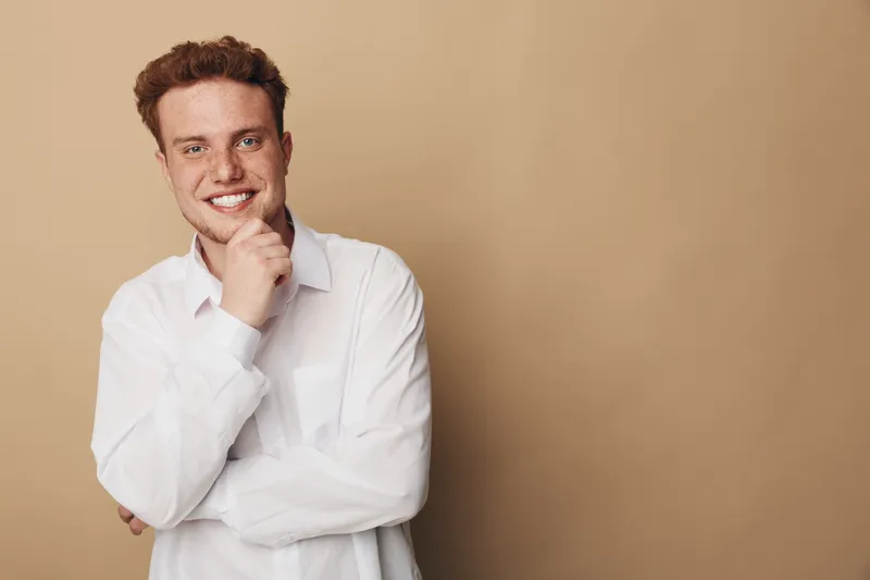 Smiling young man with curly hair wearing white shirt poses with hand on chin against beige background. Concept of confidence, casual style, and positive mood in studio portrait.