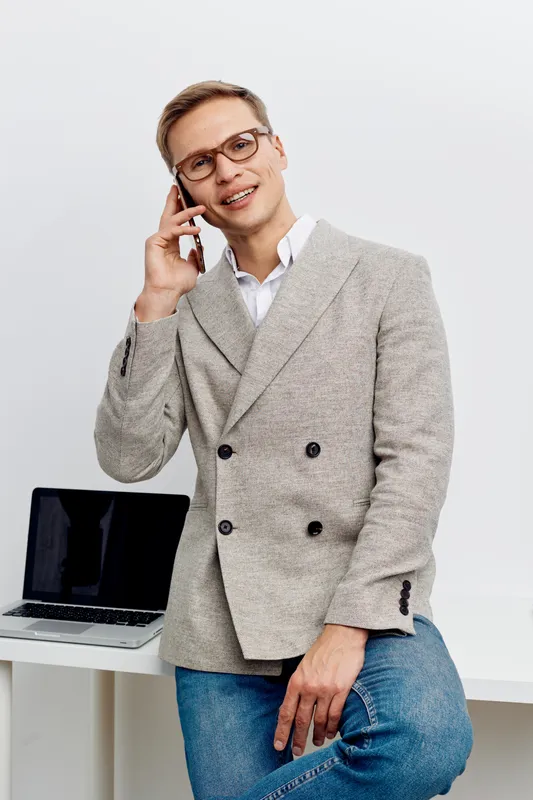 Smiling man wearing glasses talking on mobile phone in office setting, dressed in smart casual gray blazer and jeans, with laptop on white desk, light background, professional and friendly atmosphere.