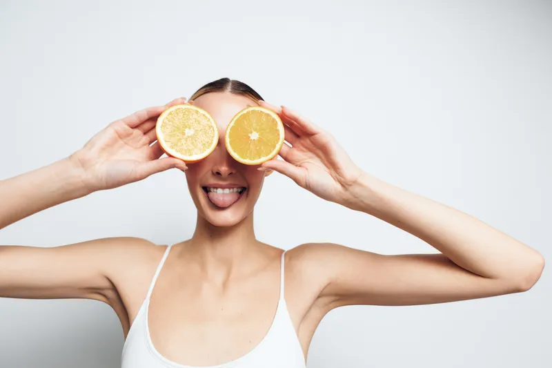 Young woman holding two halves of orange in front of eyes, playful expression, sticking out tongue, white tank top, clean background, fresh and fun mood.