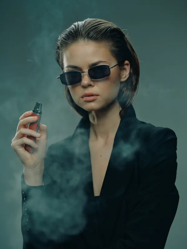 Stylish woman with sunglasses holding vape device surrounded by smoke in dark fashion concept