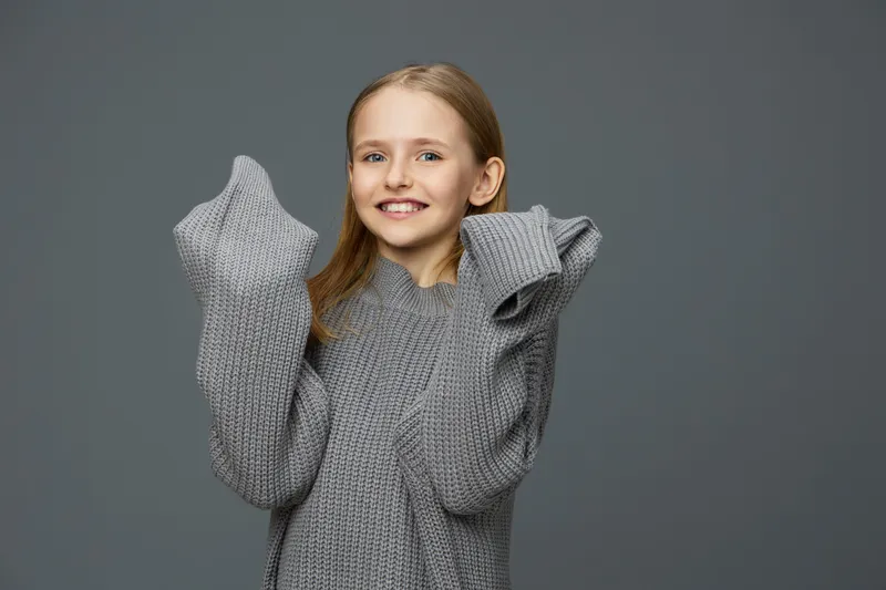 Young girl in oversized gray sweater expressing joy with a bright smile against a neutral gray background, perfect for concepts of comfort and happiness