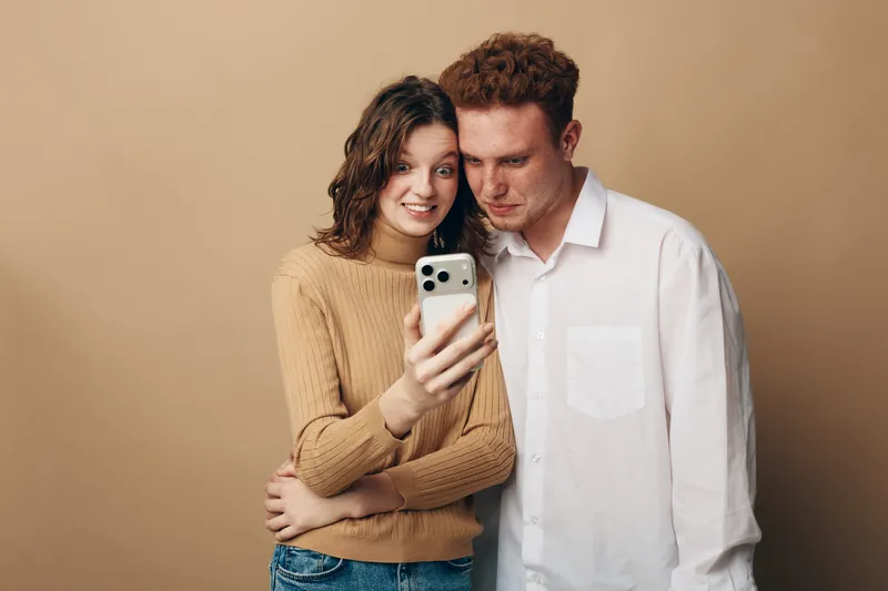 Young couple with curly hair takes selfie on smartphone. Woman in beige sweater smiles while man in white shirt looks at screen. Concept of friendship, technology, and casual lifestyle.