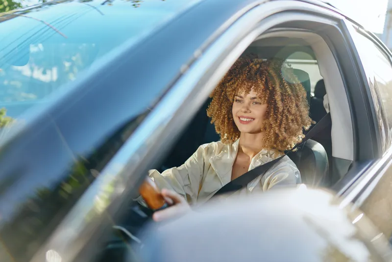 smiling woman driving car with seatbelt fastened on sunny day, casual clothing, curly hair, urban background, safe driving concept
