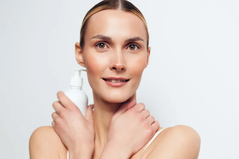 Smiling woman with clean skin holding a white lotion bottle close to her face on a white background. Concept of skincare, beauty, health, and self-care with soft natural lighting.