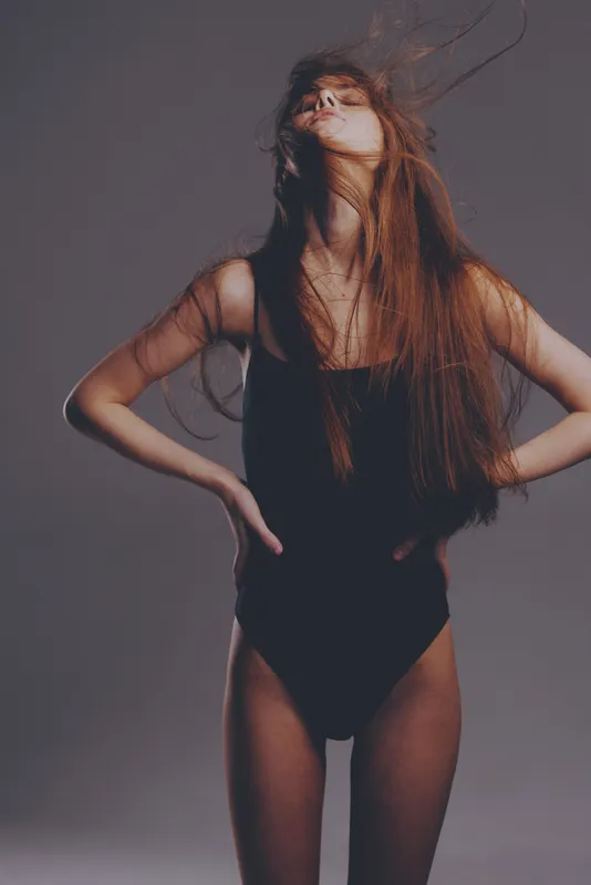 young woman in black bodysuit with flowing hair posing confidently in studio lighting