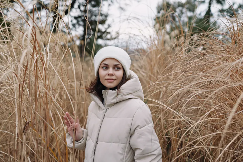 Woman in a light winter coat and white beanie stands among tall dry grass, calm expression in an outdoor wintry landscape with soft natural light and peaceful mood