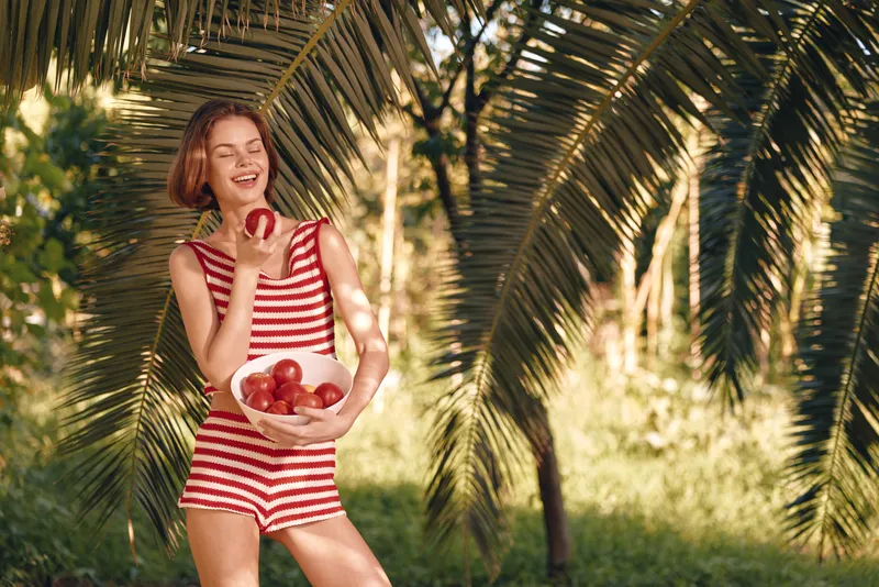 A cheerful woman in a red and white striped swimsuit enjoys a sunny outdoor setting among palm trees, holding a bowl of apples and laughing, capturing a fresh tropical mood.