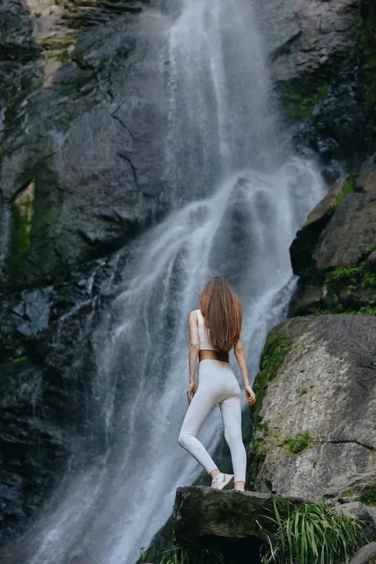 Woman hiking in white activewear standing on rock near tall waterfall surrounded by lush greenery and rugged cliffs, showcasing adventure and nature.