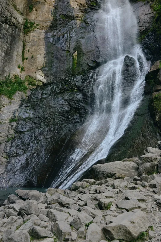 Natural waterfall cascading down rocky cliff surrounded by green moss and rugged stones under diffuse daylight in a serene mountainous landscape.