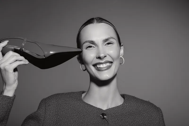 Smiling Caucasian woman with light skin holds high heel shoe near head, wearing textured sweater and hoop earrings, isolated on white background, studio portrait.