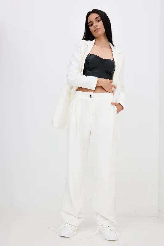 woman in white suit and black top standing confidently in minimalistic studio with neutral background, wearing white sneakers, relaxed pose, modern fashion style