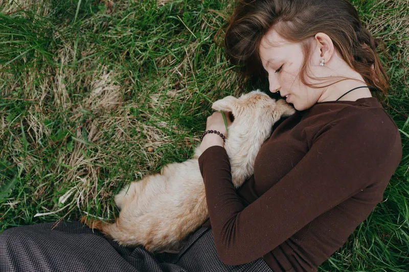 Relaxed woman outdoors, lying on grass, cuddling a gentle goat kid, warm sunlight, calm mood, soft breeze, natural colors, peaceful connection, cozy embrace, rustic scene