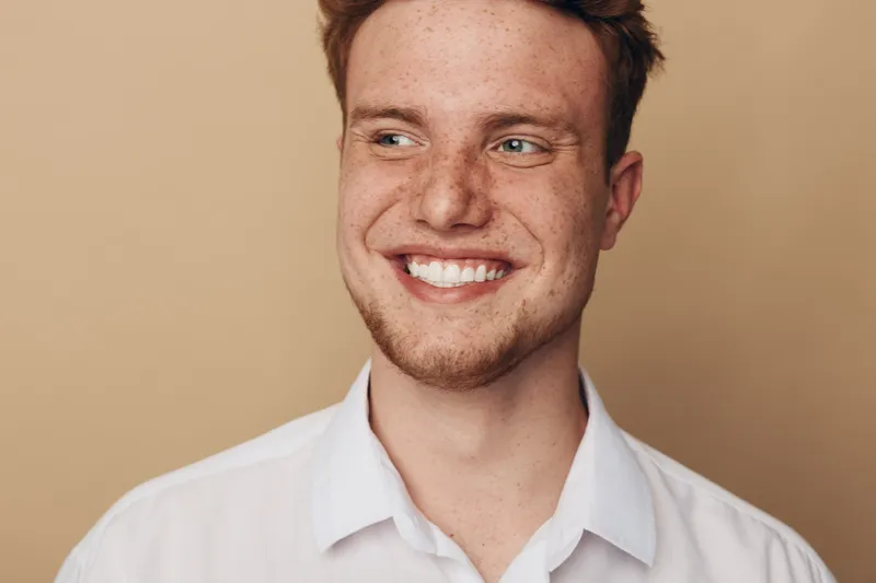 Smiling young man with freckles and short hair looks away wearing white shirt. Close-up portrait on beige background. Concept of happiness, youth, casual style, and natural beauty.