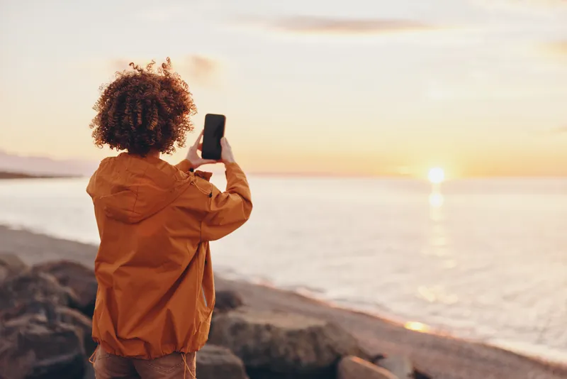 Woman lifestyle outdoors, standing on rocky beach at sunset, curly hair visible, holding phone for photography, warm golden light, casual jacket, relaxed pose, gentle breeze, scenic horizon