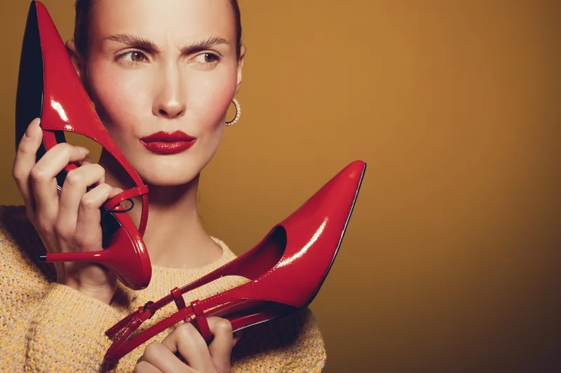 Woman with red lipstick holds red high heel shoes near face, looking away with thoughtful expression. Fashion concept with bright colors and stylish accessories.