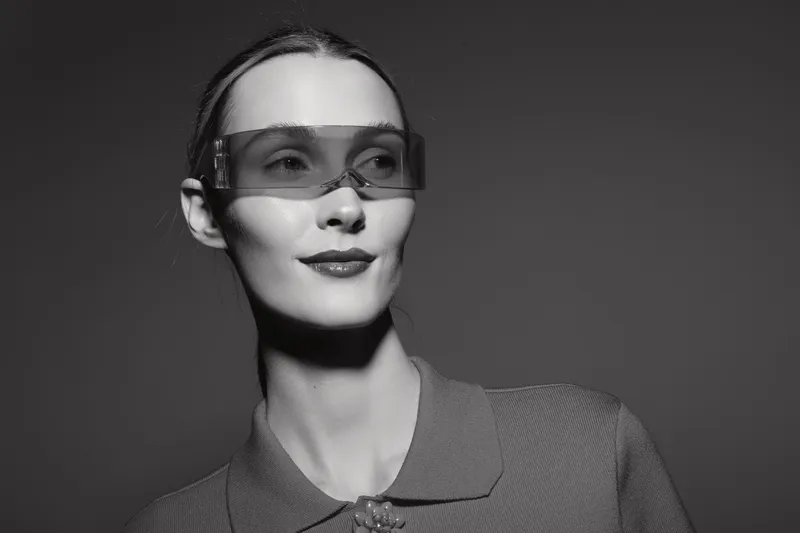 Portrait of a young white woman with slicked hair wearing futuristic sunglasses and a textured jacket in studio light.