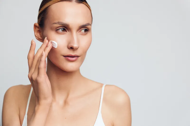 Young woman applying face cream on clean skin, skincare routine, natural beauty, soft lighting, white background, focused expression, casual wear, healthy lifestyle concept