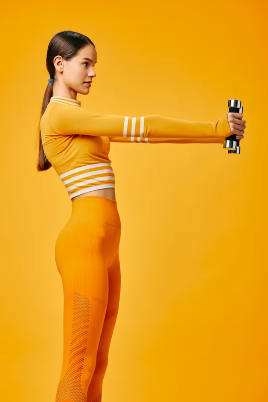 Woman fitness enthusiast in orange athletic wear performs a shoulder extension with dumbbells, dynamic exercise moment in a bold orange studio background, conveying energy, strength, and focus during