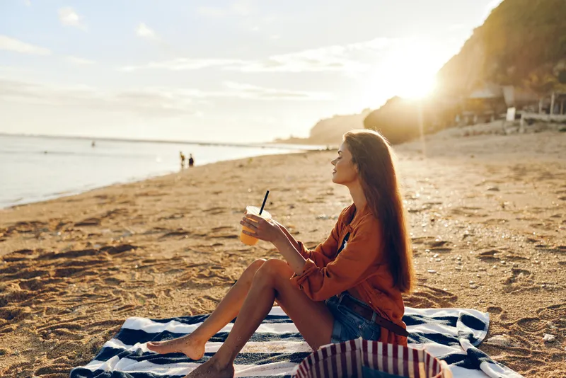 Young woman sits on a striped towel at sunset, sipping a drink and gazing toward the calm sea, a relaxed, reflective moment with warm light on her skin