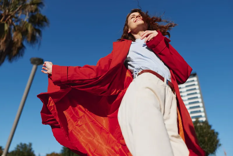 A young woman in a bright red coat twirls outdoors under a clear blue sky, her cape flowing as she smiles confidently and moves with freedom and energy.