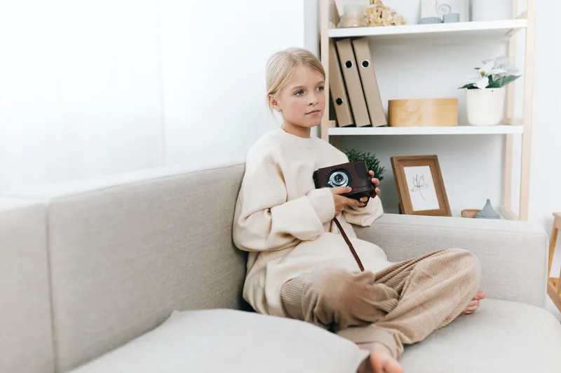 Authentic playful scene of a young student at home, camera in hand, capturing curiosity, imagination, and daily school life in a cozy living room setting.