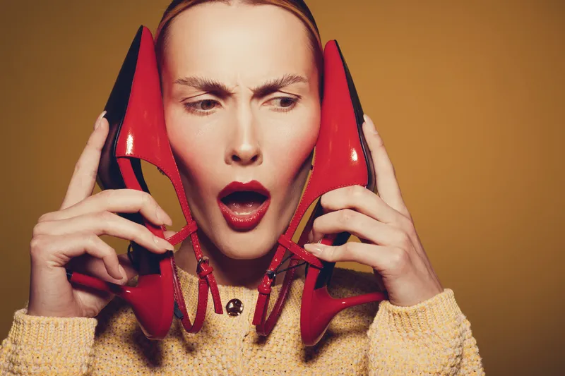 Young woman with red lipstick holds red high heel shoes near her face, expressing surprise and curiosity. Studio portrait with warm brown background and soft lighting.
