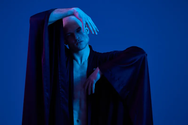 A striking image of a man posing dramatically in a dark robe against a vibrant blue background, showcasing artistic expression and contemporary fashion.