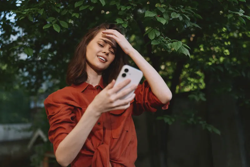 Cheerful woman outdoors, posing with smartphone, casual red shirt, sunlit garden scene, candid lifestyle moment, natural colors, leafy backdrop