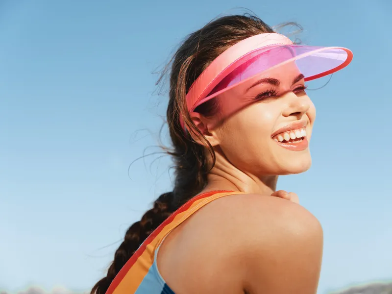Bright outdoor portrait of a joyful woman wearing a pink visor and colorful athletic top; sunlit skin and clear blue sky create a vibrant, energetic moment.