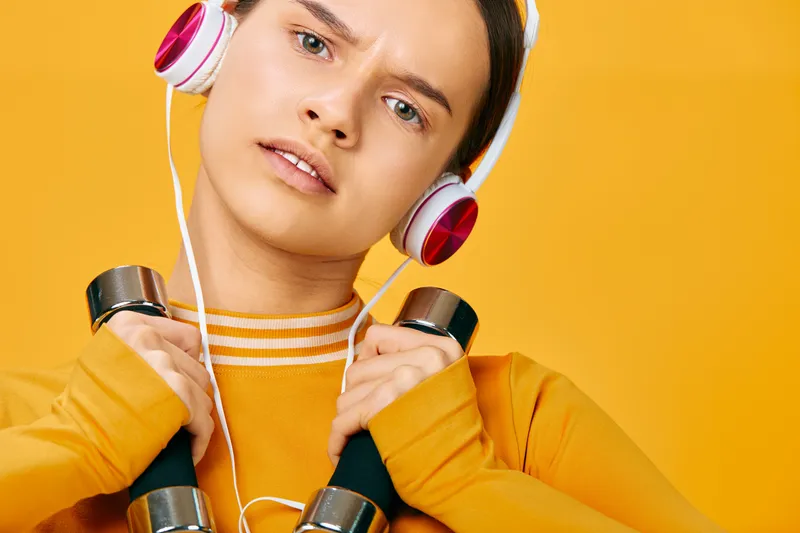 Fitness focused portrait of a woman in athleticwear lifting dumbbells with headphones against a bright orange backdrop, embodying sport, workout and energy in a dynamic studio scene.