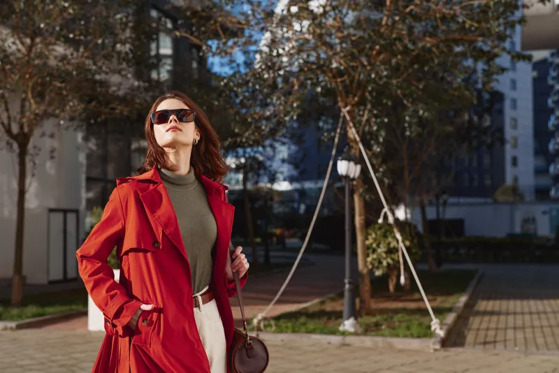 Elegant brunette woman in a red trench coat wears a green turtleneck and carries a dark bag, standing on a city street with sunglasses, exuding confident chic
