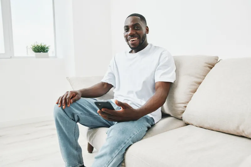 Happy African American man in casual white t shirt sitting on a sofa with smartphone, smiling and enjoying his time at home in a bright interior
