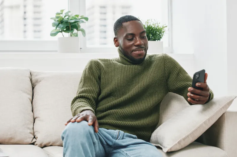 Young Black man in a cozy green sweater happily browsing on his smartphone while relaxing on a sofa in a bright, modern living room