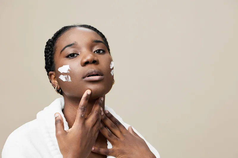 Young woman applying skincare cream on her face, with a thoughtful expression and a neutral background, emphasizing self care and beauty routines