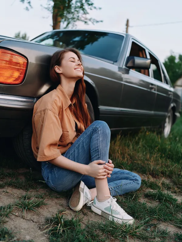 Young woman enjoying a serene moment outdoors, sitting by a vintage car, wearing casual attire and smiling in a warm, sunny atmosphere.