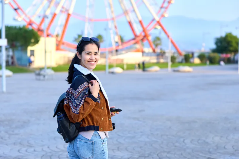 Smiling woman with a backpack poses in a sunny outdoor plaza near a ferris wheel, wearing a casual jacket and jeans for a relaxed amusement park travel moment today