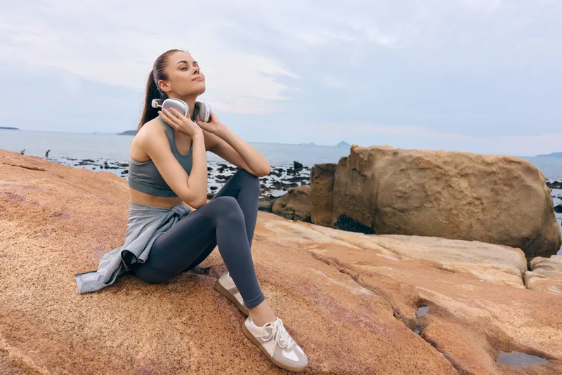 Young woman enjoying music outdoors on rocky beach, wearing comfortable athletic clothing and headphones, relaxing in a serene natural setting