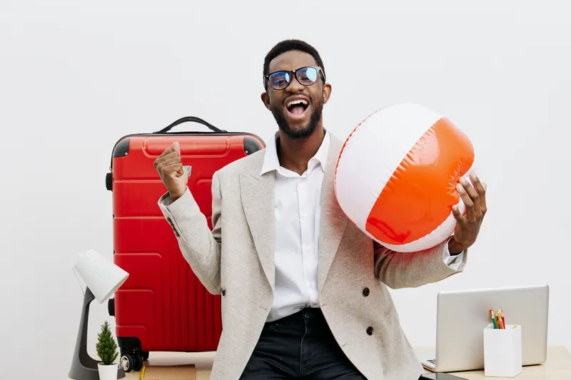 Stylish smiling young man with afro hairstyle and glasses, wearing a light blazer, holding a large beach ball in one hand and giving a thumbs up, standing in modern office with travel gear. People lif