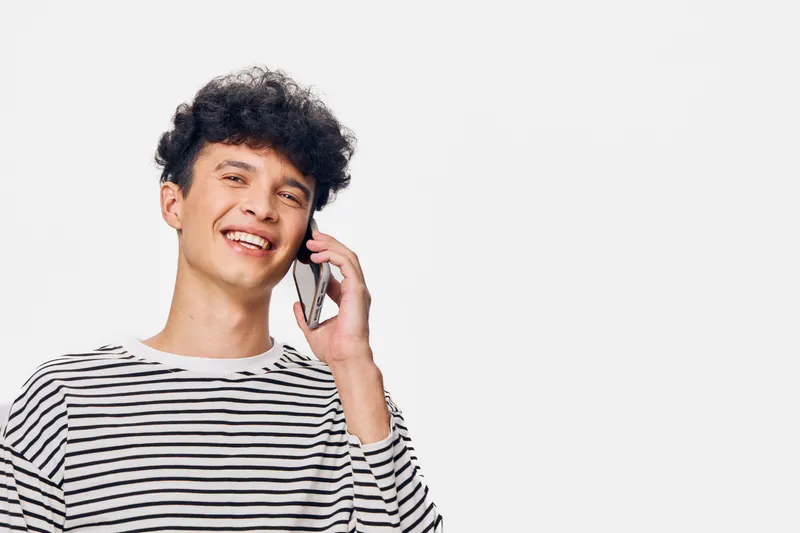smiling young man talking on smartphone wearing striped shirt against white background, casual style with cheerful expression and curly hair