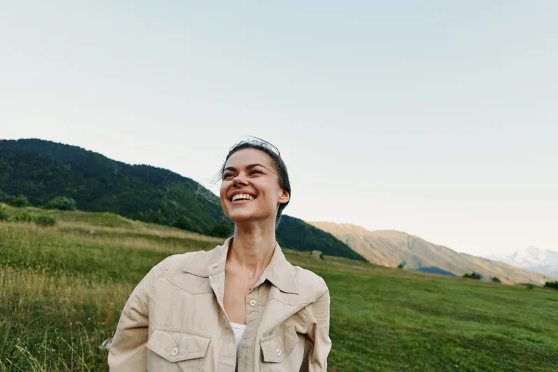 Young woman outdoors in a grassy field smiles up at the sky, enjoying fresh air and sunlit peace as distant mountains rise behind her in a warm breeze, casual pose, natural colors, sense of freedom.
