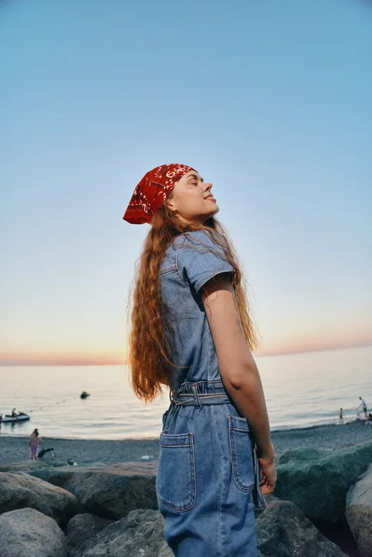 Lifestyle woman in modern boho-western denim street style enjoys sunset by the sea with film color tones, relaxed and stylish outdoors moment capturing freedom.