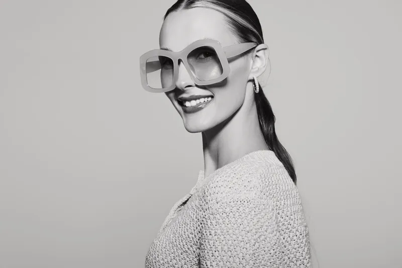 Smiling white woman with pale skin wearing oversized sunglasses and knitted sweater poses against plain background. Concept of fashion, style, confidence, and modern beauty.