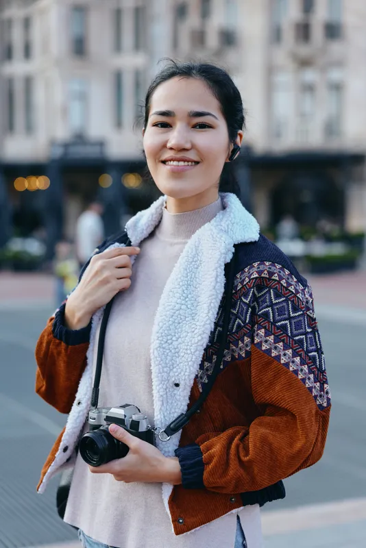 Camera in hand, smiling woman stands outdoors in a stylish jacket with a cozy fleece lining, urban plaza backdrop, evoking street photography, travel vibes and spontaneous creative moments